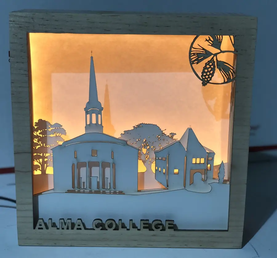 Layered laser-cut wood lightbox of a college chapel and campus, lit warm from behind